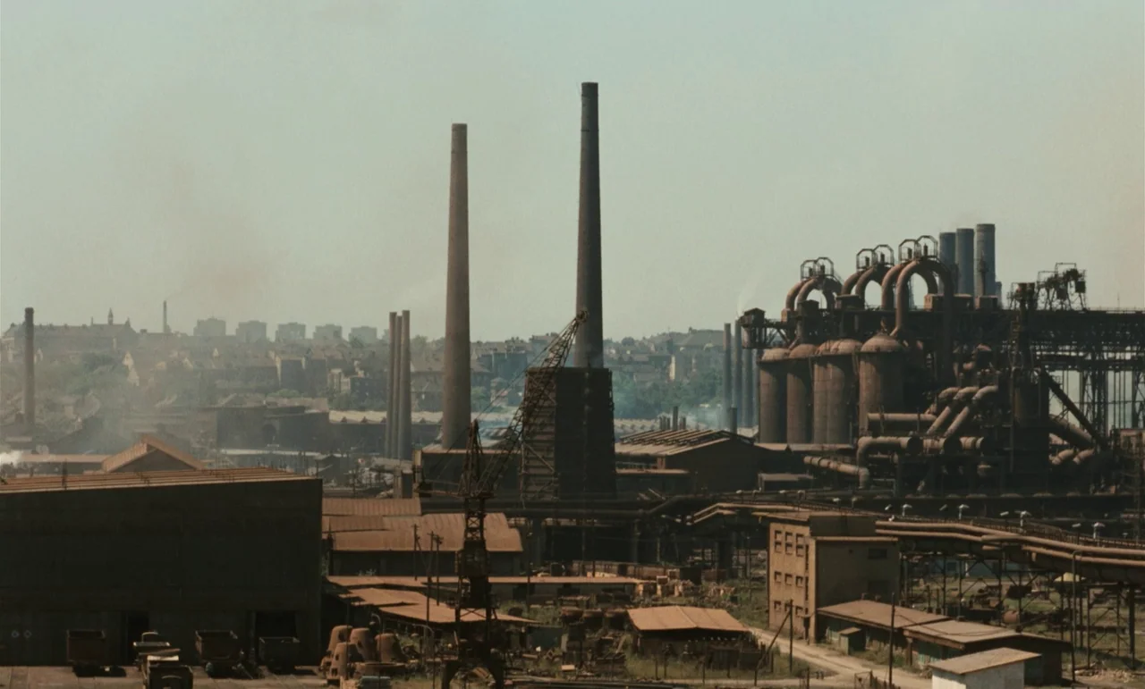 Larks On A String (1969) movie still: establishing — A vast, sprawling industrial facility with large rusted metal tanks and tall chimney stacks.…