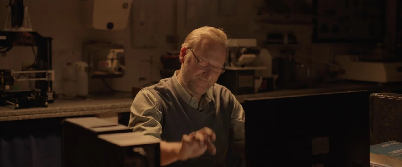 Doublespeak (2021) movie still: medium — An interior lab space features laboratory equipment on counters against a back wall. An older man sits at a…