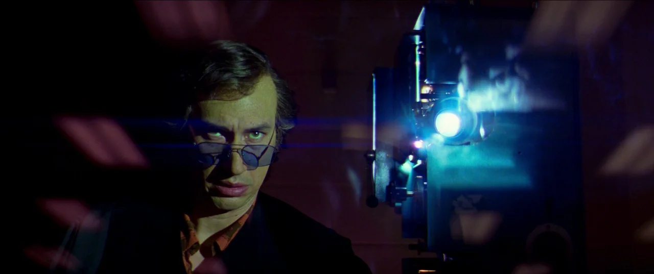 White Noise (2022) movie still: medium — A dark, cramped room contains a mechanical film projector glowing with blue light. A man wearing tinted…
