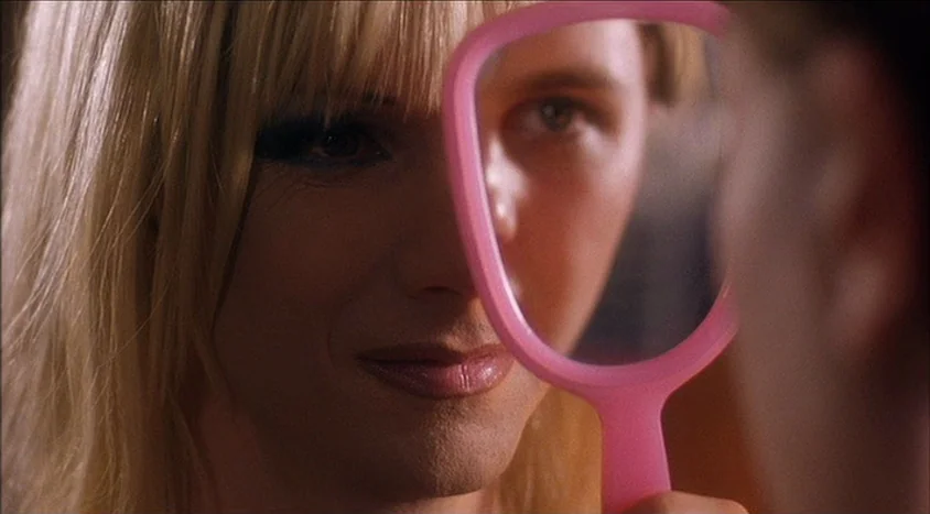 Hedwig and the Angry Inch (2001) movie still: closeup — A close-up view of a blonde woman holding a pink-framed hand mirror. She gazes directly into…