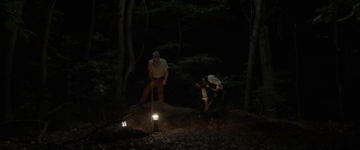 To Dust - Nightmares (2018) movie still: wide — A dense patch of dark woods with two men digging a hole at night. Two portable lanterns cast a small…