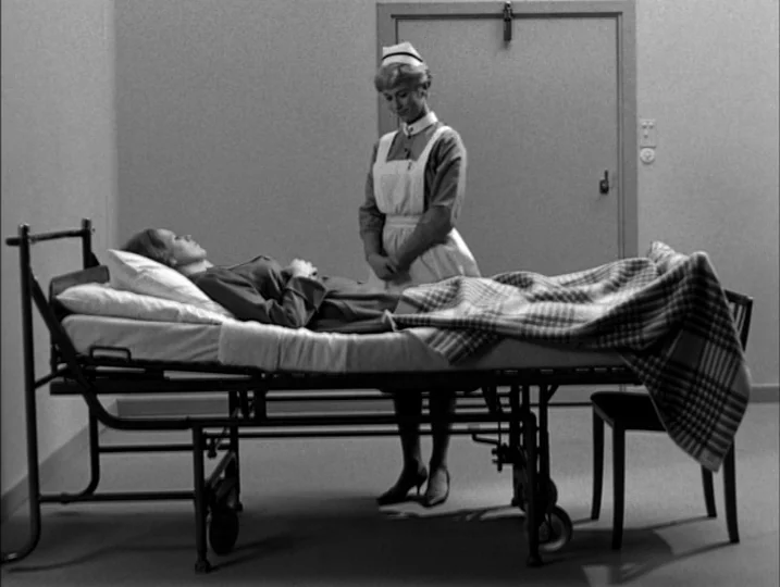 Persona (1966) movie still: wide — A sparse hospital room with plain walls and a functional metal rolling bed. A nurse stands by the bedside looking…