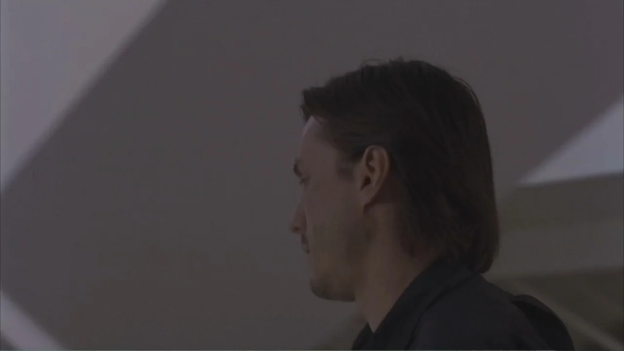 A Distant Place (2020) movie still: medium — A plain interior room with neutral walls and soft, indirect lighting. A man stands in side profile…