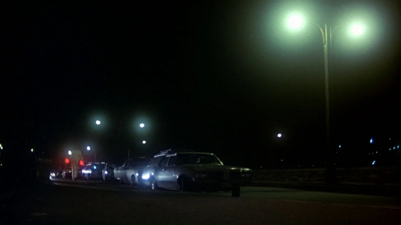 Saturday Night Fever (1977) movie still: wide — A desolate road at night is illuminated by tall, greenish streetlamps overhead. A lone figure stands…
