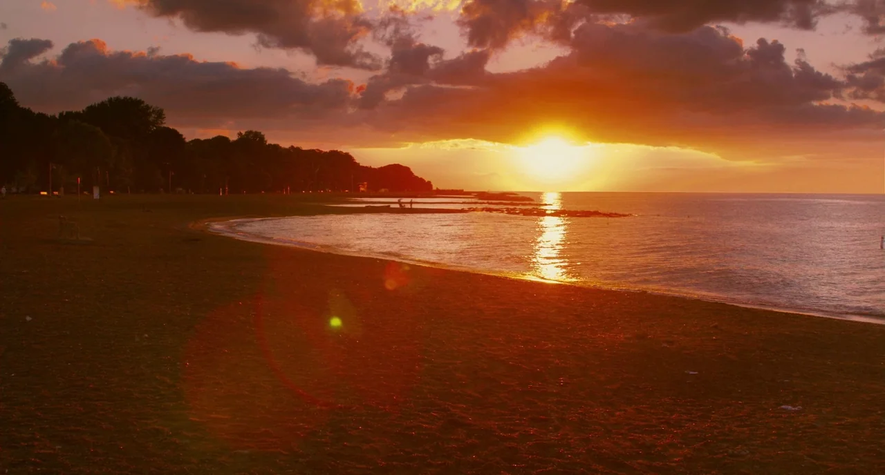 Take This Waltz (2011) movie still: establishing — A shoreline with dark sand meets a body of water under a fiery sunset. The sun sits low on the…