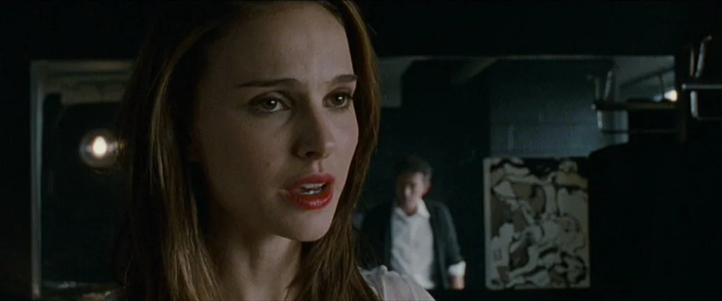 Black Swan (2010) movie still: closeup — A dark dressing room featuring a mirror reflecting a man in the background. A young woman with brown hair…