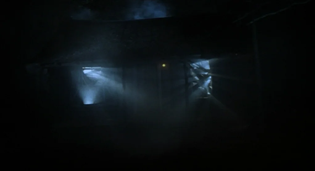 Evil Dead II (1987) movie still: establishing — A dark, dilapidated wooden cabin is obscured by thick fog at night. Beams of bright white light…