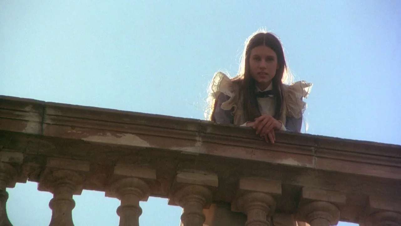 Picnic at Hanging Rock (1975) movie still: medium — A stone balustrade stands against a bright, clear blue sky during the day. A young woman in a…