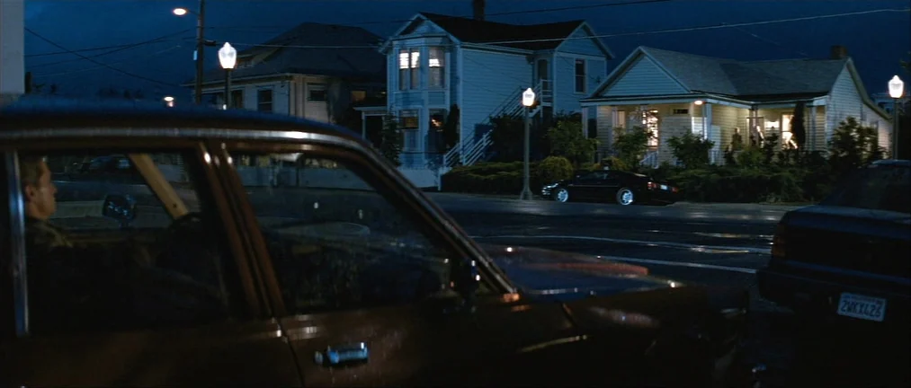 Basic Instinct (1992) movie still: medium — A suburban street at night features quiet houses and parked cars under electric light. A man sits inside…