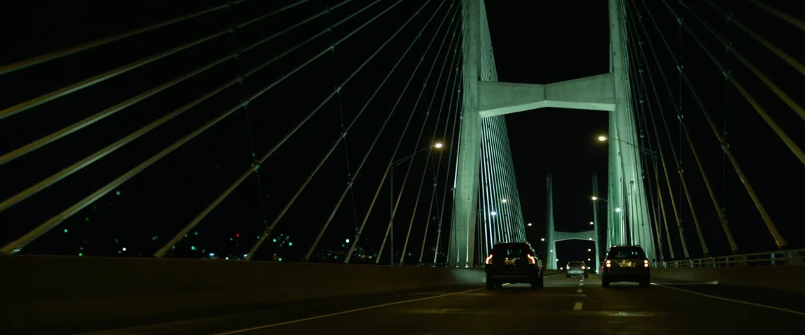 Gone Girl (2014) movie still: wide — A large cable-stayed bridge spans the darkness under a night sky at night. Several vehicles travel across the…
