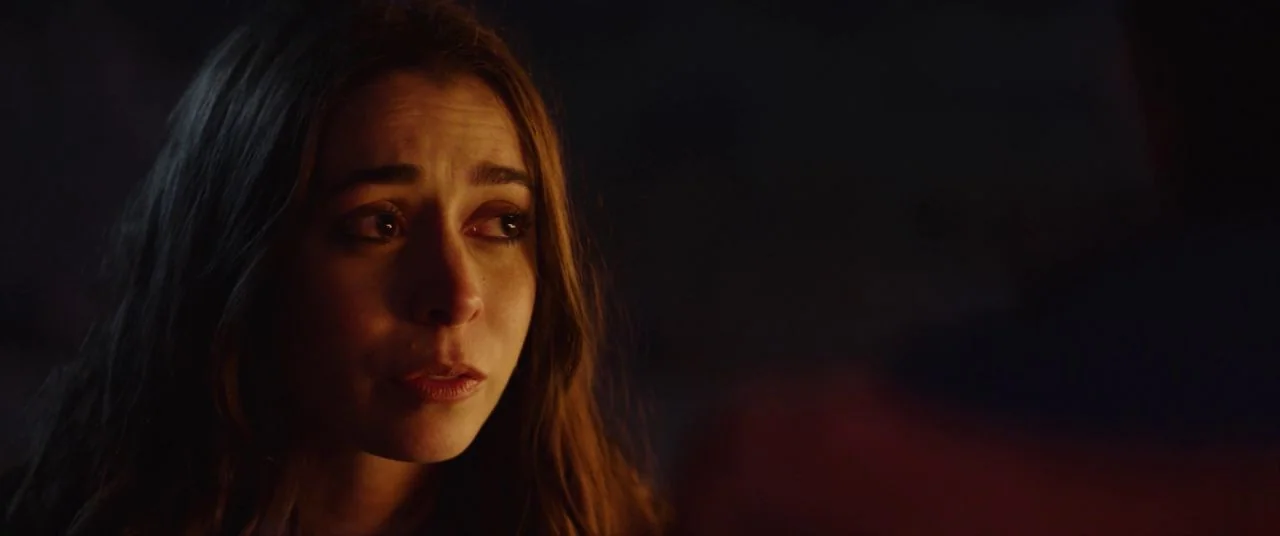 Palm Springs (2020) movie still: closeup — A dark outdoor space illuminated by a nearby fire at night. A young woman with brown hair stares intently…