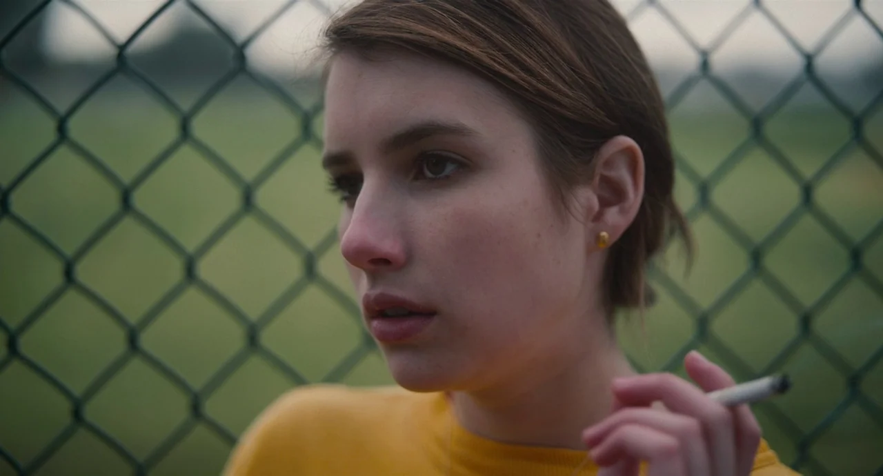 Palo Alto (2013) movie still: closeup — A woman stands in front of a green chain link fence. She holds a lit cigarette while looking toward the left…