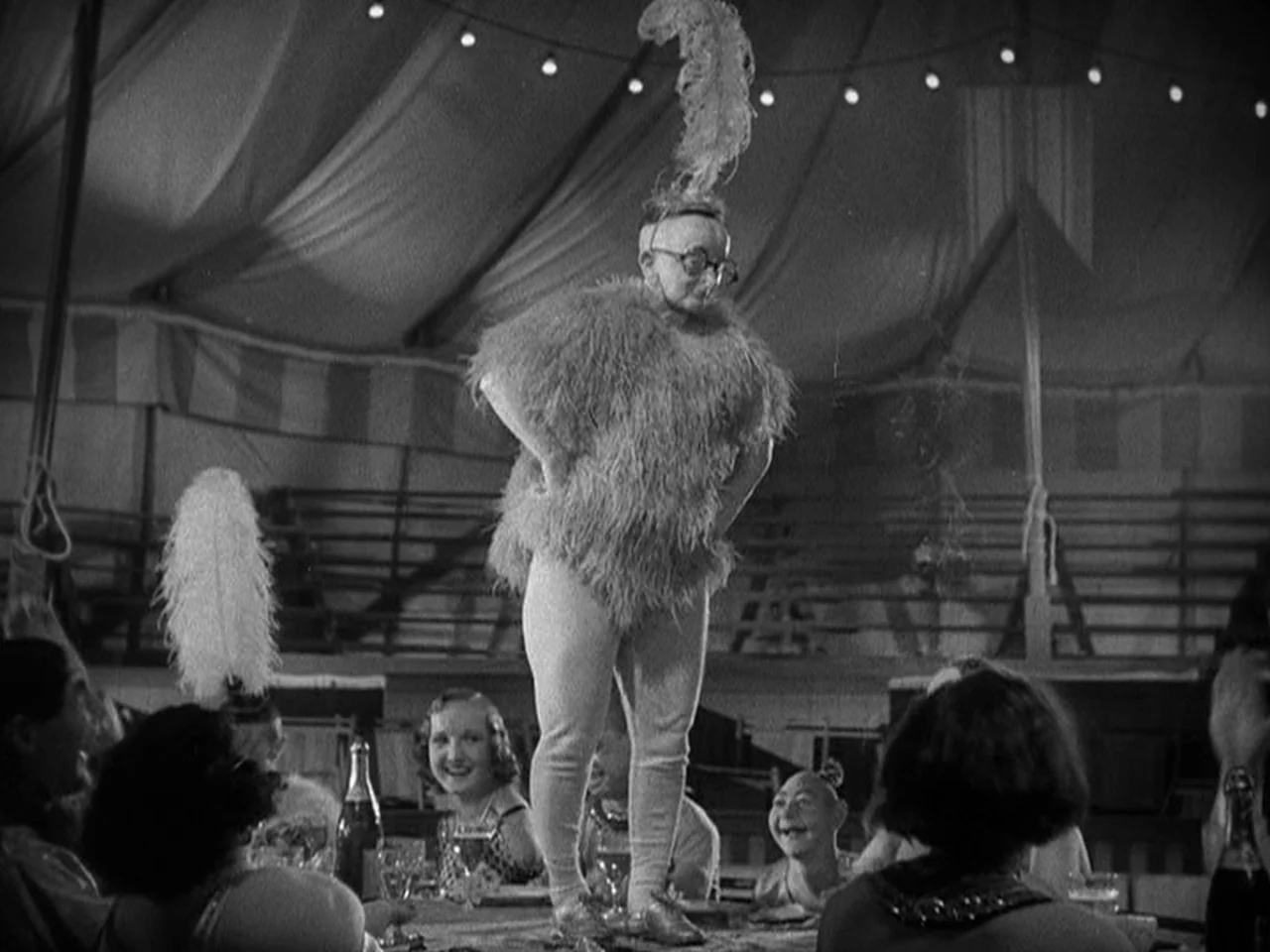 Freaks (1932) movie still: fullbody — The interior of a large circus tent features draped fabric and rows of seating. A person in a feathered…