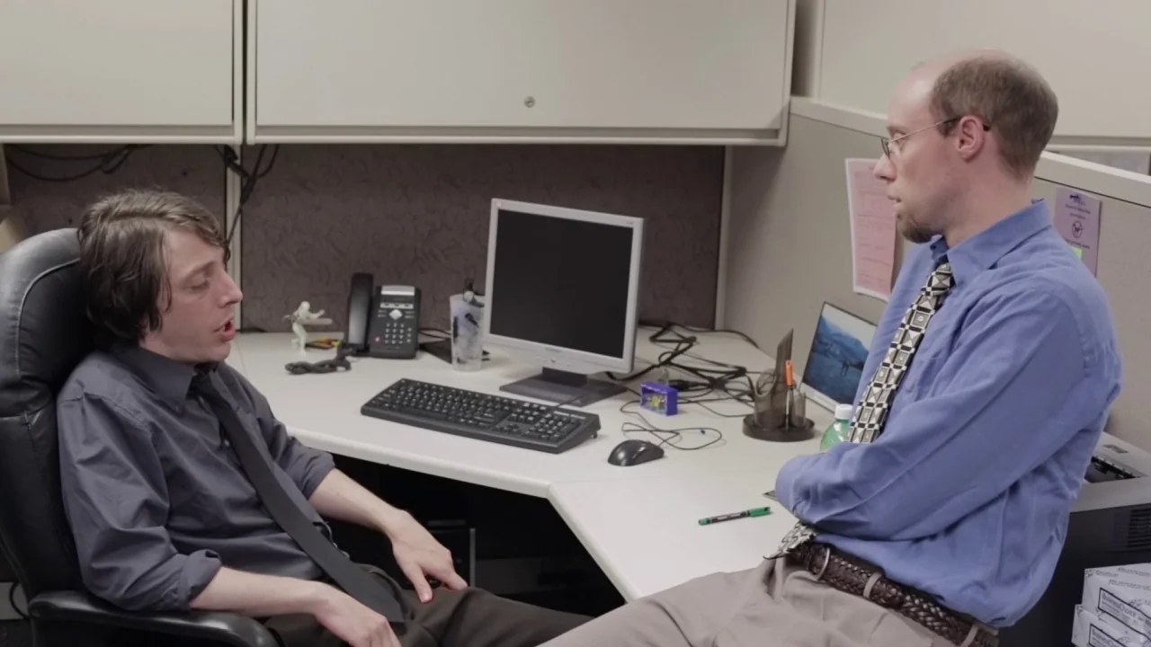 Buzzard (2014) movie still: medium — A cramped corporate cubicle features a desk with a computer, phone, and office supplies. Two men in formal…