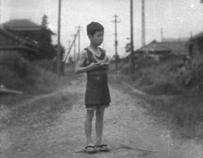 Tokyo Chorus (1931) movie still: fullbody — A rural dirt road passes between simple wooden structures and utility poles. A young boy stands alone in…