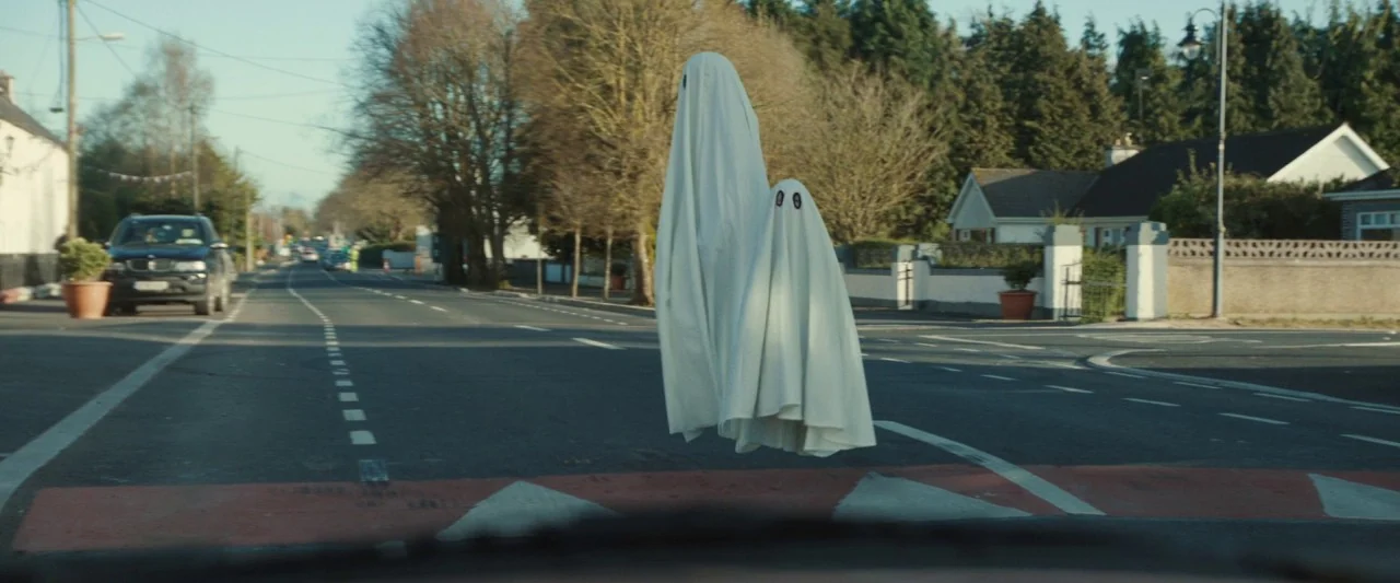 Extra Ordinary (2019) movie still: wide — A quiet suburban road intersection features residential houses and trees in the background. Two people…