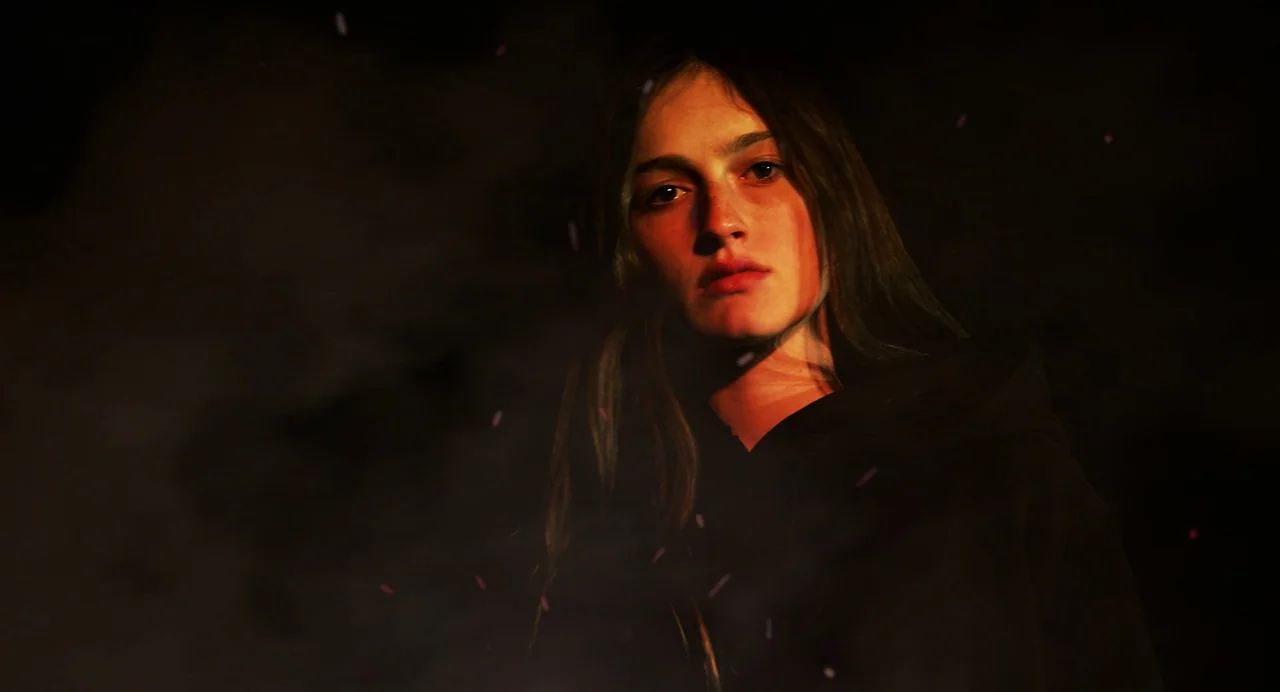 Hellbender (2021) movie still: closeup — A woman stands in darkness surrounded by faint drifting sparks. She looks forward while light casts orange…