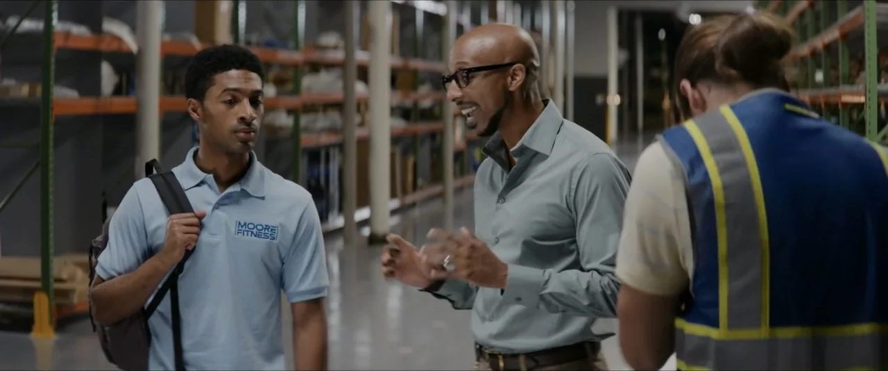 The Forge (2024) movie still: medium — An industrial facility features rows of metal shelving stocked with boxes. Three men converse in a central…