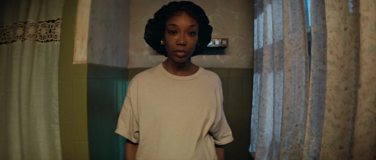 The Front Room (2024) movie still: medium — A small room features sage green tiled walls and a window with patterned curtains. A woman in a white…
