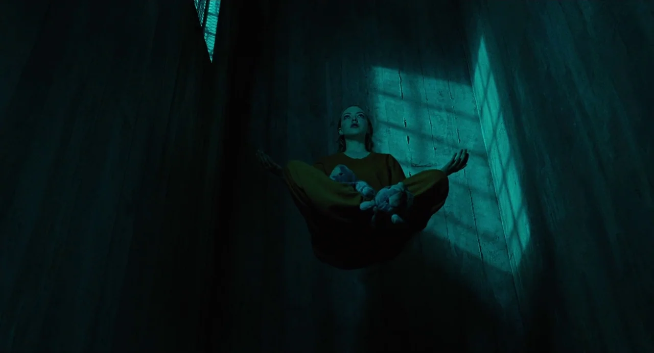 Jennifer’s Body (2009) movie still: wide — A dark concrete-walled room features a high, barred window casting a sharp light shape. A woman levitates…