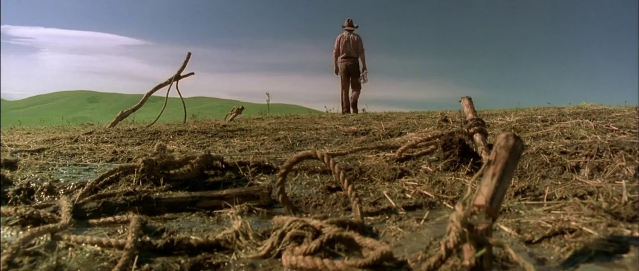 Open Range (2003) movie still: wide — An open rural field features muddy ground littered with abandoned ropes and wooden stakes. A lone man in…