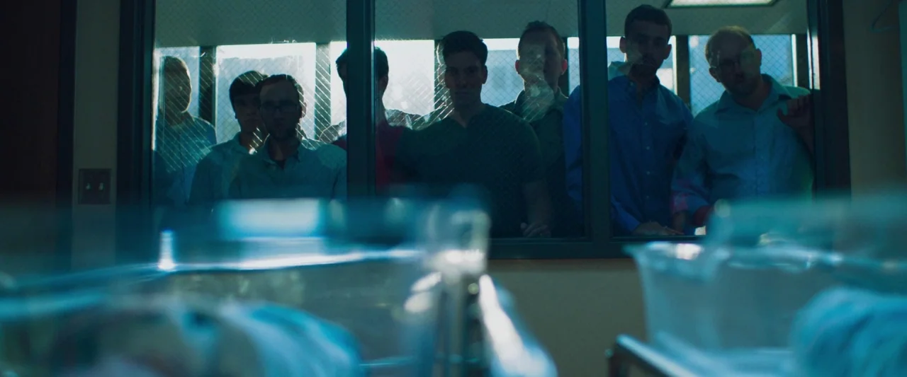 Mom and Dad (2017) movie still: wide — A row of men stand behind a wired glass window inside a brightly lit room. They look forward toward two…
