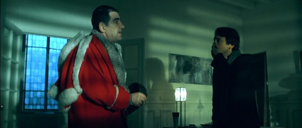Mortal Transfer (2001) movie still: medium — A dimly lit room with walls cast in long, striped shadows from window blinds. Two men stand facing each…