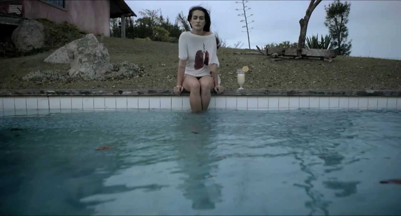 Suspension Points (2022) movie still: medium — A woman sits on the edge of an outdoor swimming pool during the day. She wears a white graphic…