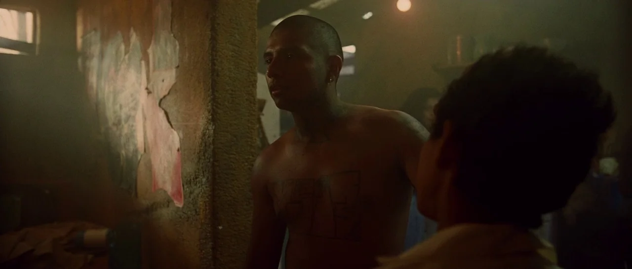 Sin Nombre (2009) movie still: medium — A dimly lit room features walls with peeling, textured surfaces in a hazy atmosphere. An shirtless, tattooed…