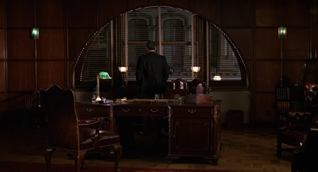 Miller’s Crossing (1990) movie still: wide — A dark paneled room features a large arched window behind an ornate mahogany desk. A man stands at the…
