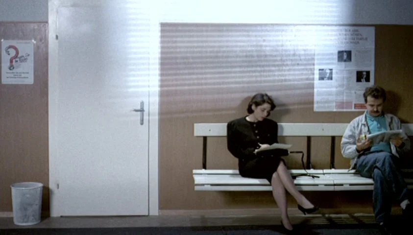 The Seventh Continent (1989) movie still: wide — An interior waiting area features a long white bench positioned against a paneled wall. A woman and…