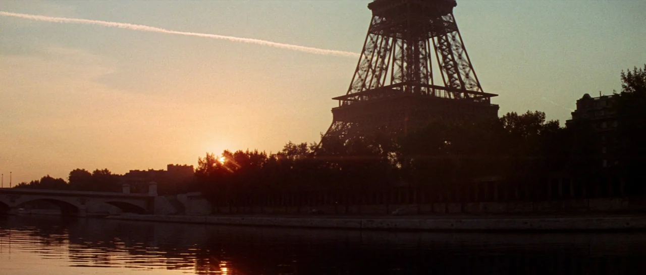 Irma La Douce (1963) movie still: wide — The Eiffel Tower silhouettes against a sunset sky over a calm riverbank. A stone bridge stretches across…