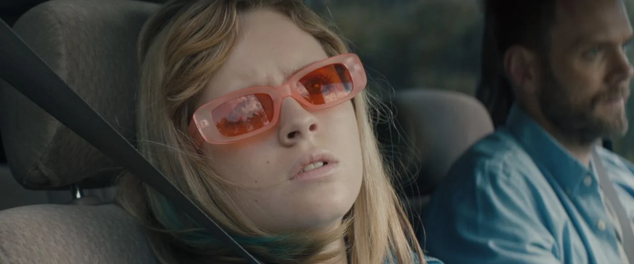 Becky (2020) movie still: medium — The interior of a moving car during daylight hours with blurred scenery outside. A woman in pink sunglasses sits…