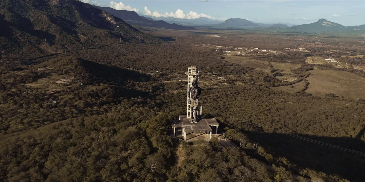 Leyenda Viva (2022) movie still: establishing — A tall concrete statue stands on a platform overlooking a vast hilly landscape. The statue is…