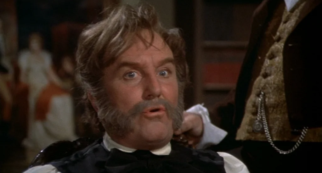 Demons of the Mind (1972) movie still: closeup — A dimly lit room with out-of-focus figures visible in the background. A man with distinct facial…