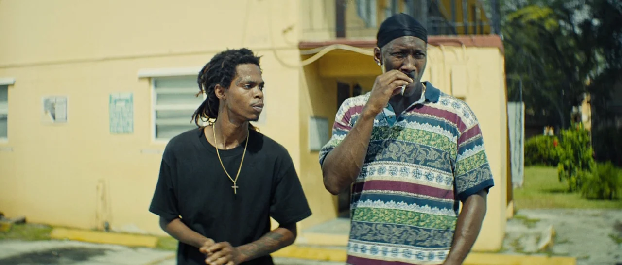 Moonlight (2016) movie still: medium — The exterior of a yellow stucco apartment building features a porch and parking area. Two men stand in front…