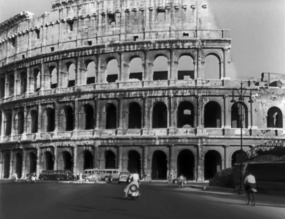Roman Holiday (1953) movie still: establishing — The Colosseum stands prominently against a bright sky, its weathered stone arches showing history.…