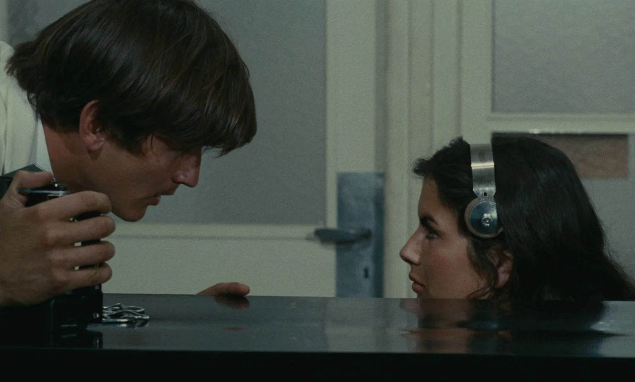 Z (1969) movie still: medium — A plain interior with a pale door and a dark, glossy counter surface. A man holding a camera looks down at a woman…