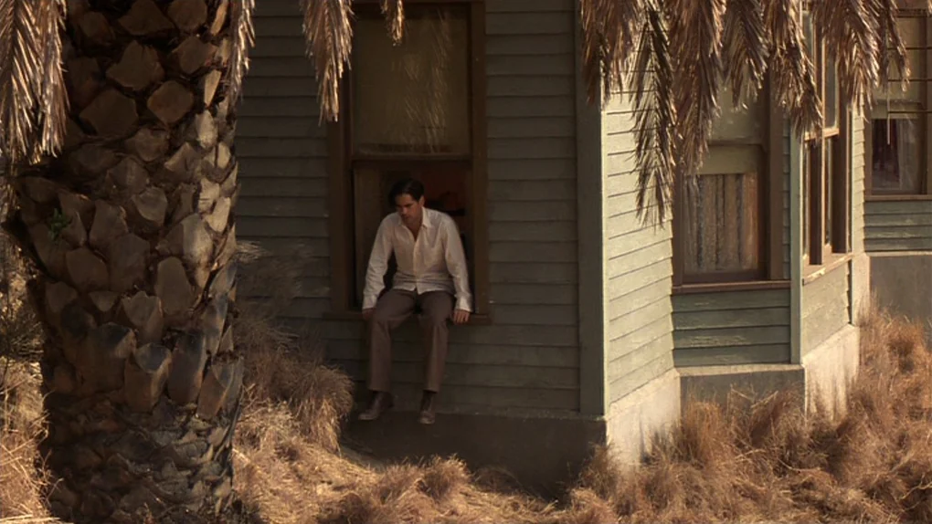 Ask The Dust (2006) movie still: wide — A wooden clapboard house sits amidst dry vegetation under a tall palm tree. A man sits motionless in the…