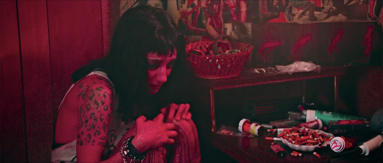 68 Kill (2017) movie still: medium — A cluttered bedside table sits in a dimly lit, red-saturated room. A woman with dark hair sits hunched over…