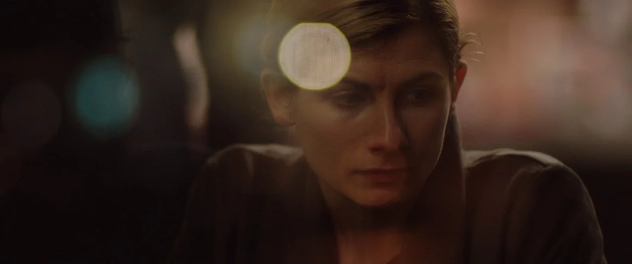 Journeyman (2017) movie still: closeup — A dark, blurred interior space with indistinct shapes and glowing bokeh circles. A woman with tied-back…