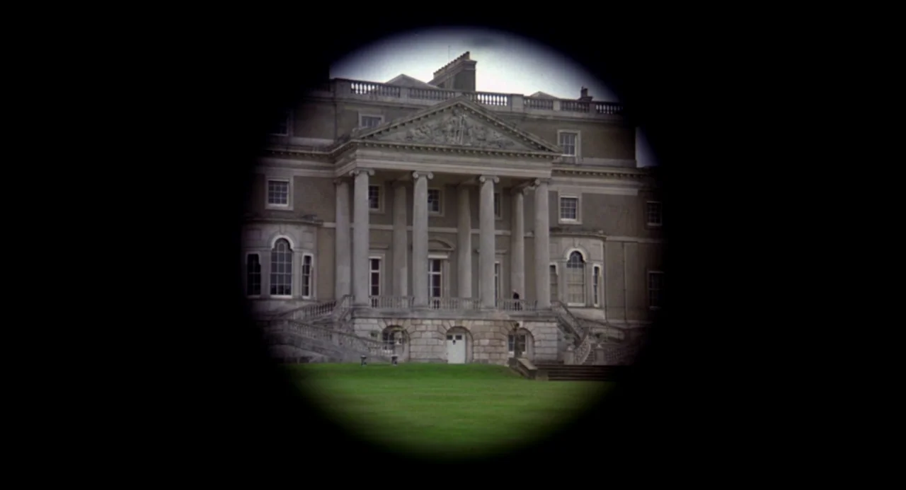 Gothic (1986) movie still: establishing — A large stone mansion with classical columns stands behind a manicured green lawn. The view is framed by a…