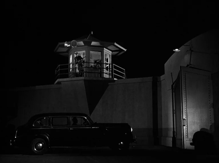 White Heat (1949) movie still: wide — A high concrete wall with a guard tower looms over an armored entrance gate. A black sedan is parked in the…