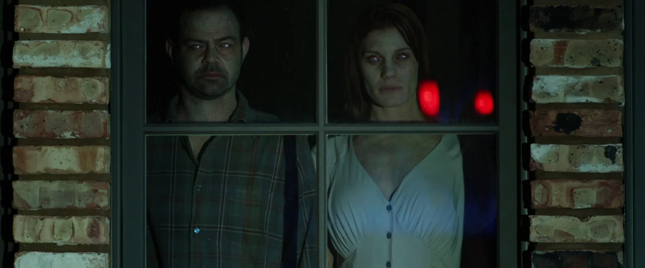 Oculus (2013) movie still: medium — A man and woman appear behind a window pane framed by exterior brick masonry. They stand motionless looking…