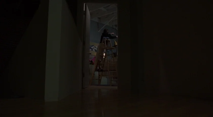 Being John Malkovich (1999) movie still: wide — A darkened hallway leads to an illuminated room viewed through an open doorway. A person stands on a…