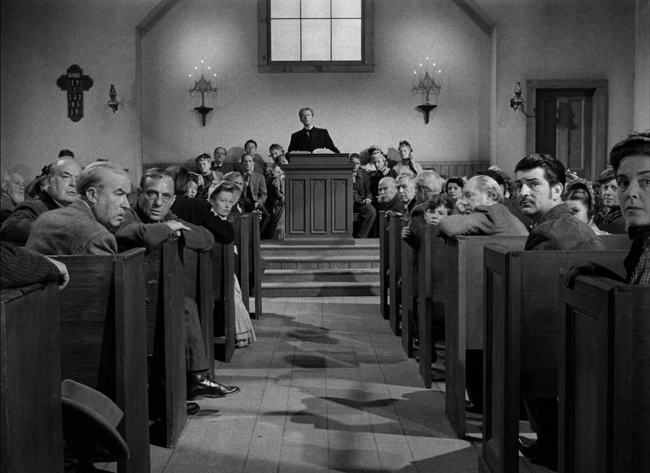 High Noon (1952) movie still: wide — A small wooden church with tiered rows of pews facing a front pulpit. A preacher stands at the podium…