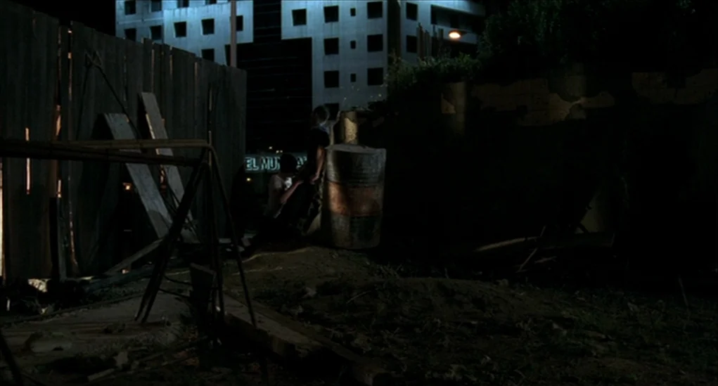 Anatomy of Hell (2004) movie still: wide — A nighttime vacant lot featuring a wooden fence and a rusted metal barrel. Two men stand together in the…