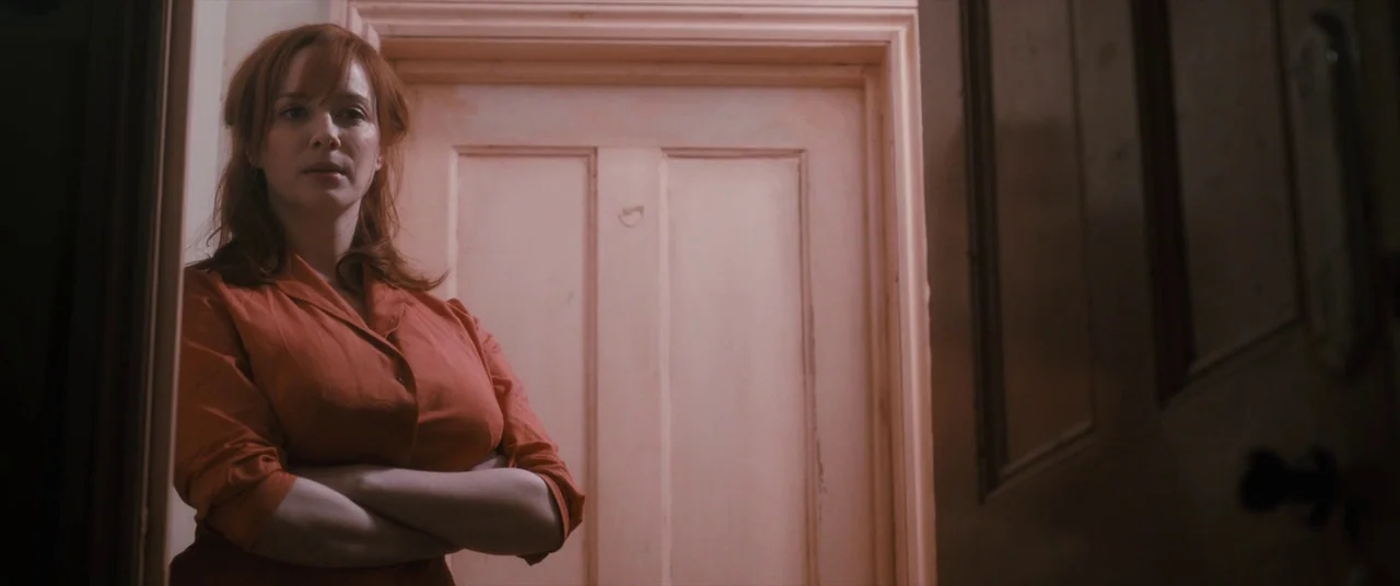 Ginger & Rosa (2012) movie still: medium — A dimly lit residential hallway features a light-colored wooden door. A woman with red hair stands in the…