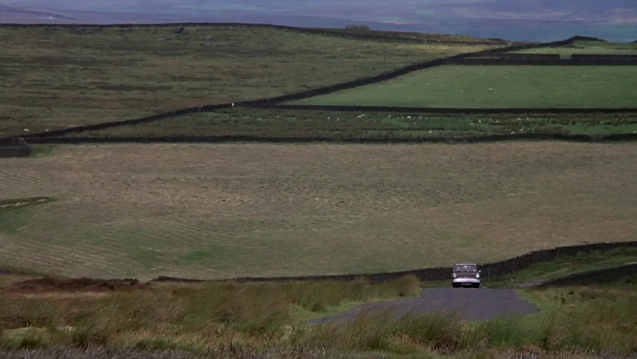 O Lucky Man! (1973) movie still: establishing — A vast, hilly landscape is divided by dry stone walls into green pastures. A lone vintage car drives…