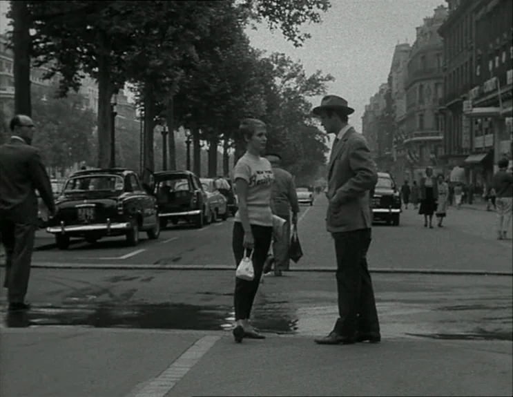 À Bout De Souffle (Breathless) (1960) movie still: fullbody — A busy Parisian street corner in the 1960s lined with parked cars and pedestrians. A…